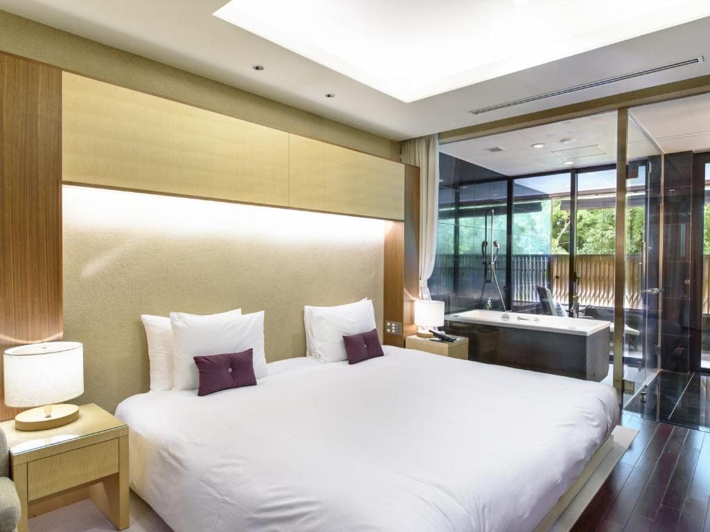 A modern hotel room with a large bed, two bedside tables with lamps, and a glass wall separating the bedroom from a bathroom with a bathtub and outdoor view. The room has warm lighting and minimalist decor.