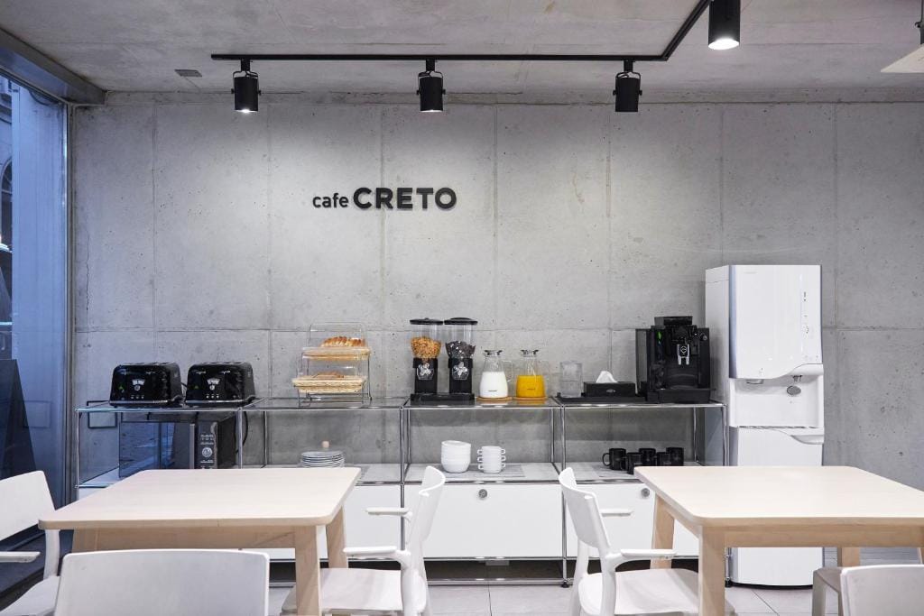 Minimalist cafe interior with light wood tables and white chairs. Against a concrete wall is a counter with toasters, snacks, drink dispensers, and coffee machines. cafe CRETO is written on the wall above.