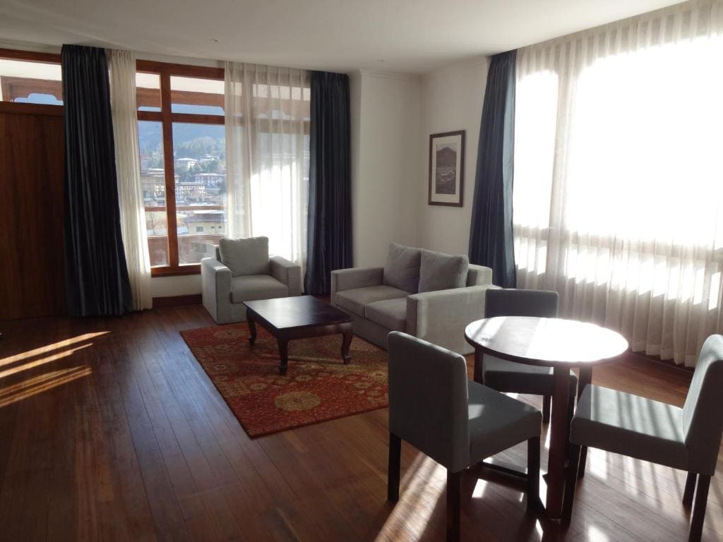 A bright living room with large windows, sheer curtains, a gray sofa set, a wooden coffee table on a patterned rug, and a small round dining table with chairs on a wooden floor.