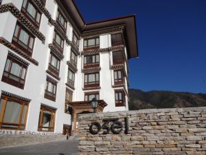 A white multi-story building with traditional Bhutanese architectural details stands under a clear blue sky. In the foreground, a stone wall features the name Osel in black letters. Trees and hills are visible in the background.