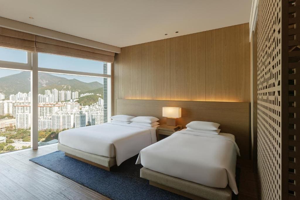 Modern hotel room with two double beds, a wooden accent wall, a nightstand with a lamp, large floor-to-ceiling windows, and a cityscape with mountains visible outside. Natural light fills the room.