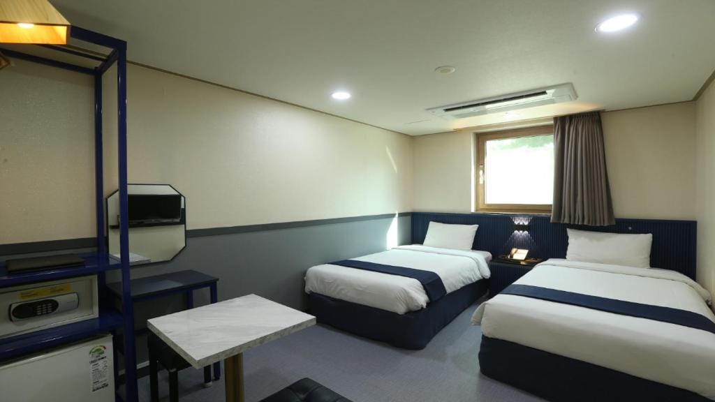 A hotel room with two single beds, a small table, a mini fridge, a safe, a wall-mounted TV, and a window with curtains. The room has neutral-colored walls and simple, modern furnishings.