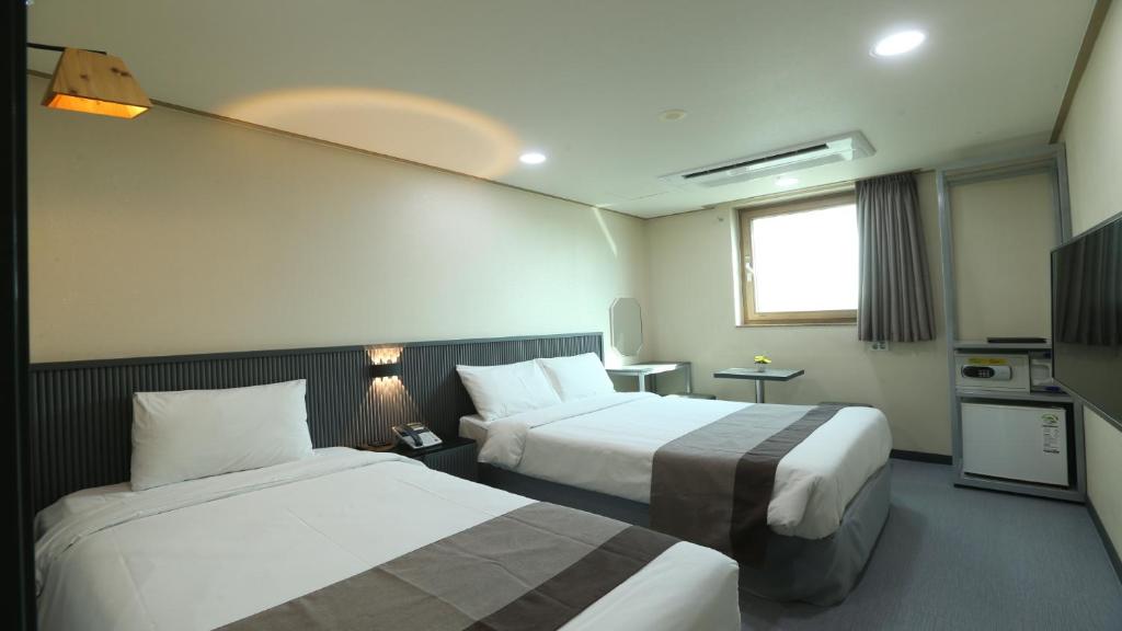 A modern hotel room with two double beds, white bedding, gray throws, a wall-mounted TV, a mini fridge, a safe, a window with closed curtains, and a small desk with a mirror and chair.