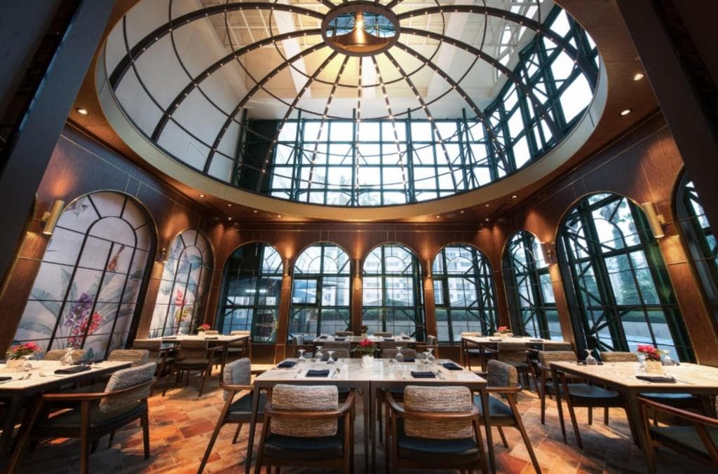 A spacious, elegant restaurant with large arched windows, a glass dome ceiling, wooden furniture, and tables set for dining, creating a bright and inviting atmosphere.