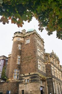 A historic stone building with Stirling Highland Hotel written on its side, featuring tall windows and a turret, framed by green leaves at the top of the image.