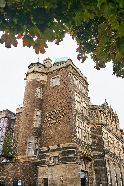 A historic stone building with Stirling Highland Hotel written on its side, featuring tall windows and a turret, framed by green leaves at the top of the image.