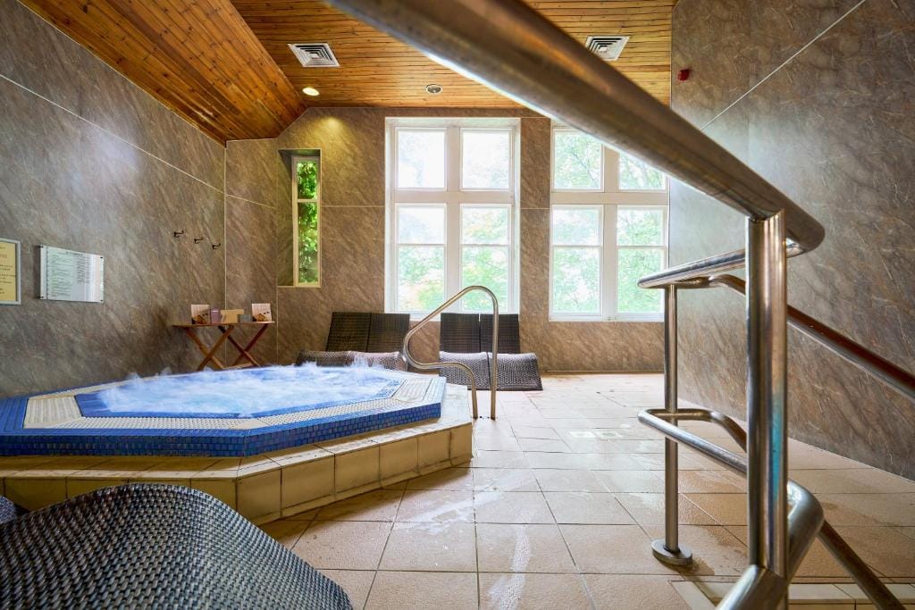 Indoor spa room with a bubbling hot tub, tile floors, lounge chairs, large windows letting in natural light, and a wooden ceiling. A metal handrail leads down to the hot tub.