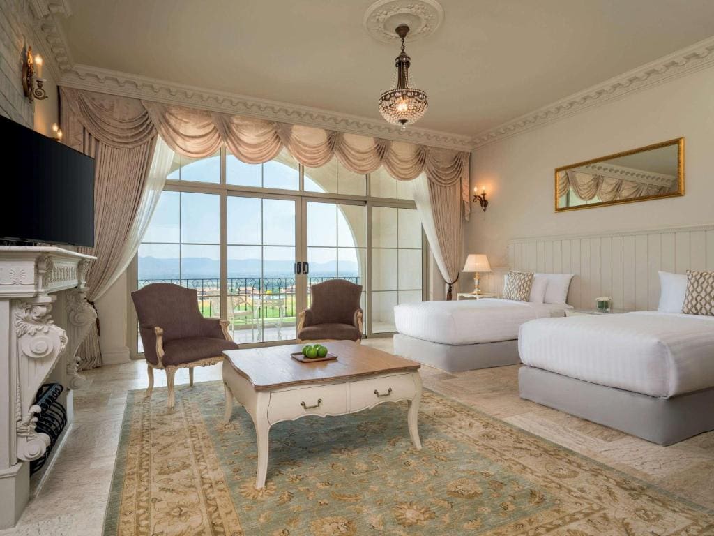 Elegant hotel room with two double beds, vintage armchairs, a coffee table, ornate chandelier, large area rug, TV, and large windows with curtains offering a view of greenery and water outside.