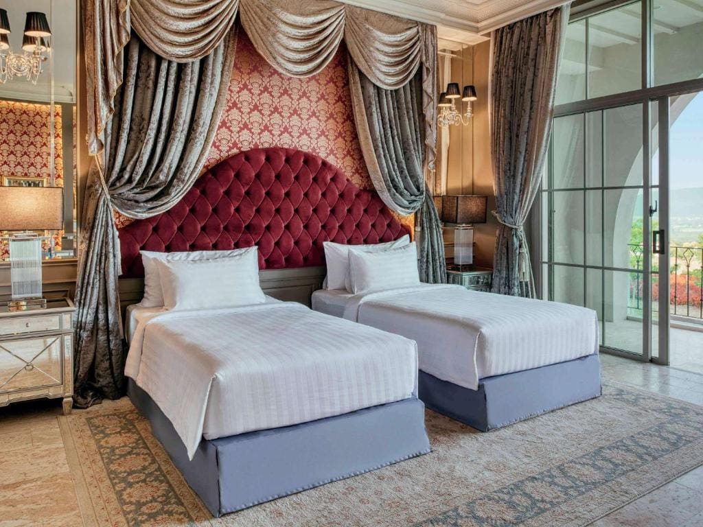 Elegant hotel room with two single beds, a large red tufted headboard, ornate drapes, mirrored nightstands with lamps, and a floor-to-ceiling window offering a scenic view.