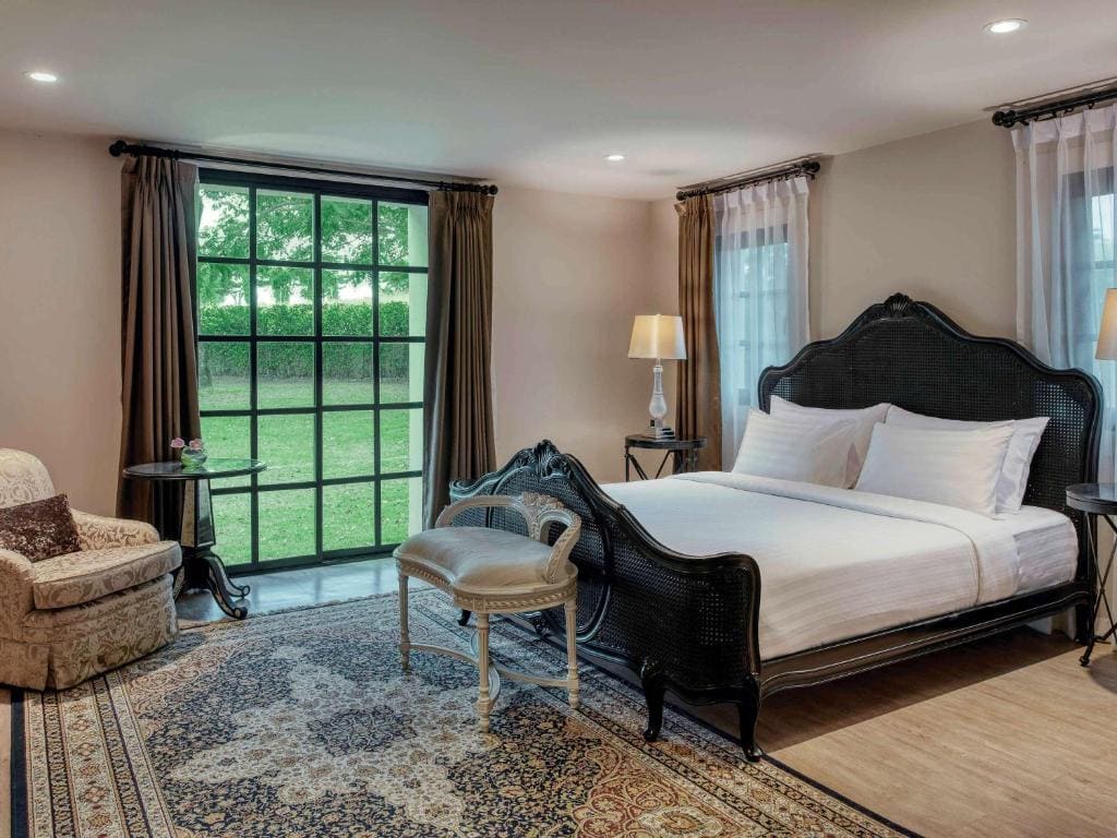 Elegant bedroom with a large black bed frame, white bedding, a patterned rug, an upholstered armchair, a cushioned bench, and large windows with sheer curtains overlooking a green lawn.