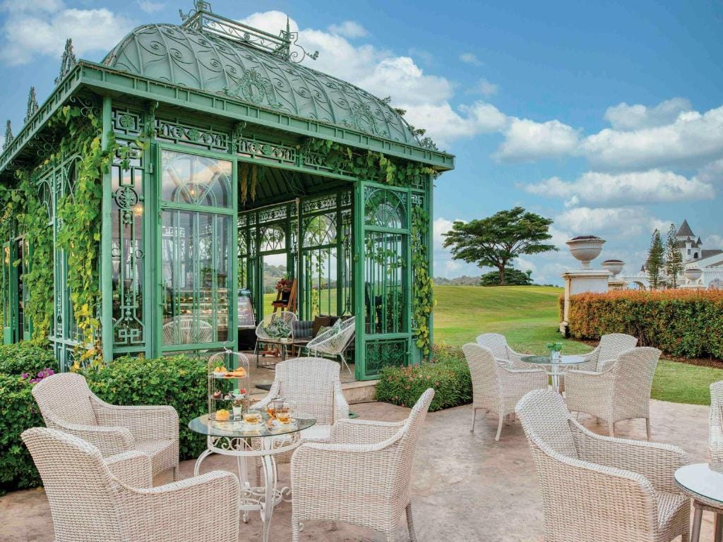 A lush garden setting with white wicker chairs and tables arranged near a green, ornate glass gazebo covered in vines, with tea and snacks served on the tables and a scenic lawn in the background.