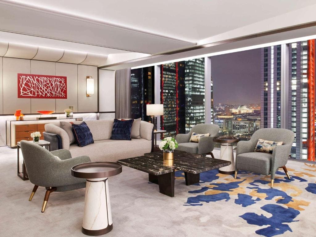 A modern, elegant living room with gray sofas and armchairs, marble tables, abstract wall art, and large windows offering a cityscape view of illuminated skyscrapers at night.