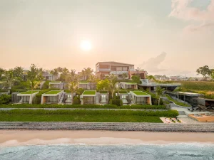 Modern beachfront resort with green rooftop villas, palm trees, and a large main building, overlooking a sandy beach and calm ocean under a warm, hazy sunset sky.