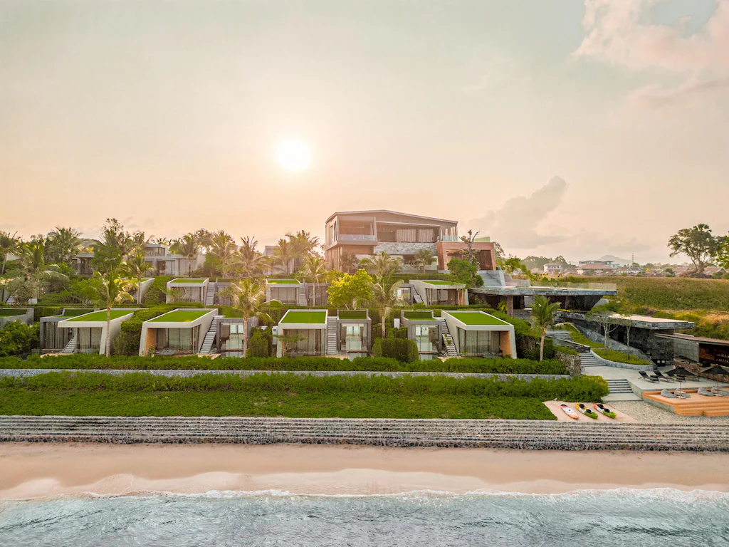 Modern beachfront resort with green rooftop villas, palm trees, and a large main building, overlooking a sandy beach and calm ocean under a warm, hazy sunset sky.