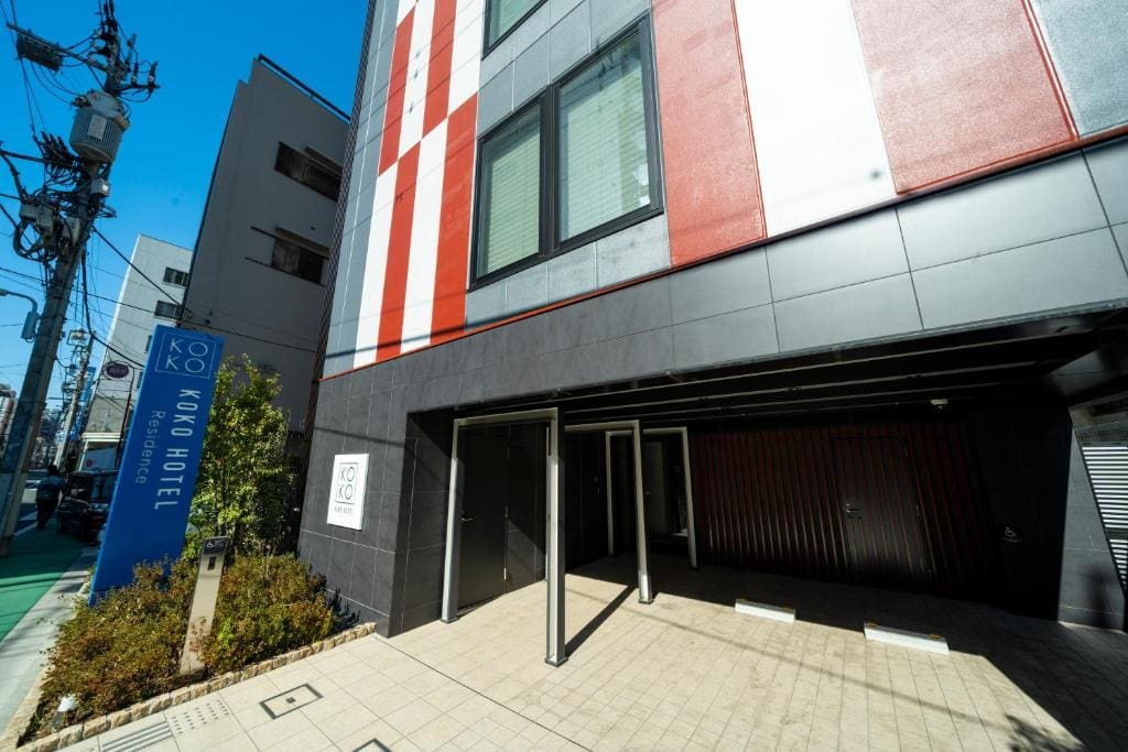 Modern hotel exterior with a vertical red, white, and gray striped design. A blue sign reads KOKO HOTEL. There is a driveway, some greenery, and utility poles visible against a clear blue sky.