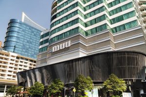 A modern, multi-story building with green-tinted windows and muii signage on its facade, surrounded by other high-rise buildings and street-level trees under a clear blue sky.