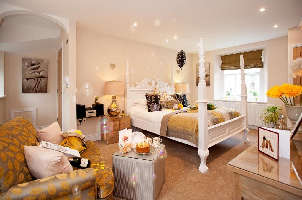 A bright, elegant bedroom with a white four-poster bed, patterned armchair, fresh flowers, a tray with tea and snacks, soft lighting, and decorative accents in gold and beige tones.