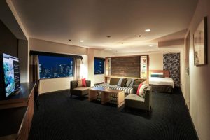 Spacious hotel suite with a large bed, sitting area with sofas and coffee table, TV, and city skyline visible through wide windows at night. Warm lighting creates a cozy atmosphere.