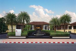 The image shows the entrance of a Marriott Resort with a central fountain, neatly trimmed hedges, palm trees, and a grand building with a sign reading JW Marriott Khao Lak Resort & Spa. A white sign stands by the driveway.