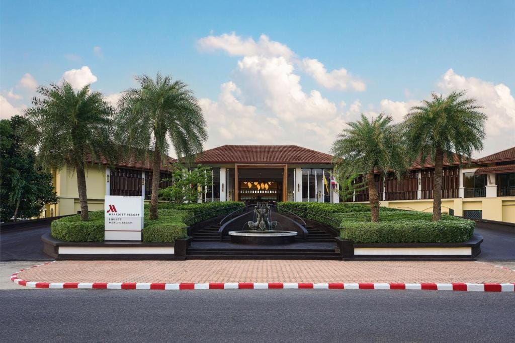 The image shows the entrance of a Marriott Resort with a central fountain, neatly trimmed hedges, palm trees, and a grand building with a sign reading JW Marriott Khao Lak Resort & Spa. A white sign stands by the driveway.