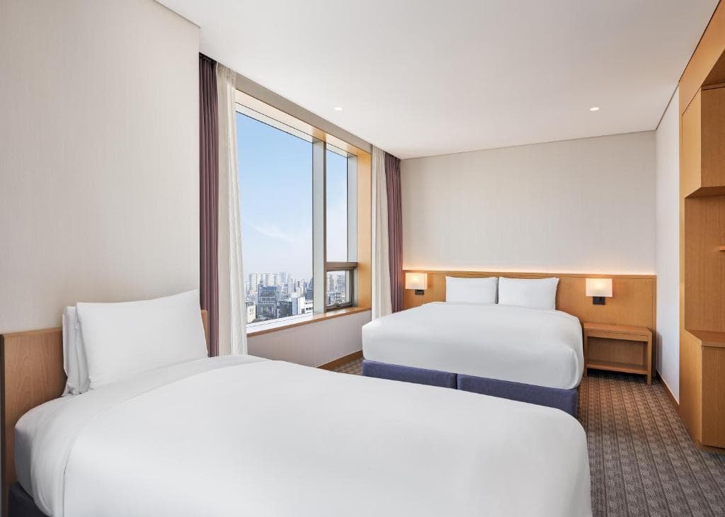 A hotel room with two beds, white bedding, a large window with city views, bedside tables with lamps, and light-colored walls and furniture.