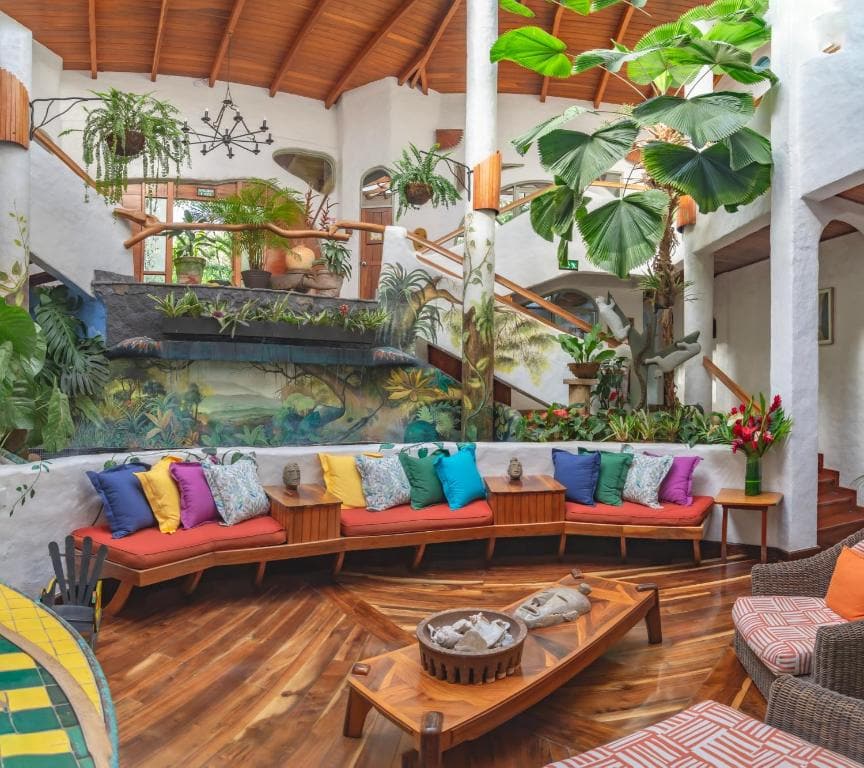 A bright, tropical living room with a curved wooden sofa covered in colorful pillows, lush indoor plants, wooden floors, a coffee table, and a tall staircase with greenery and natural light from large windows.