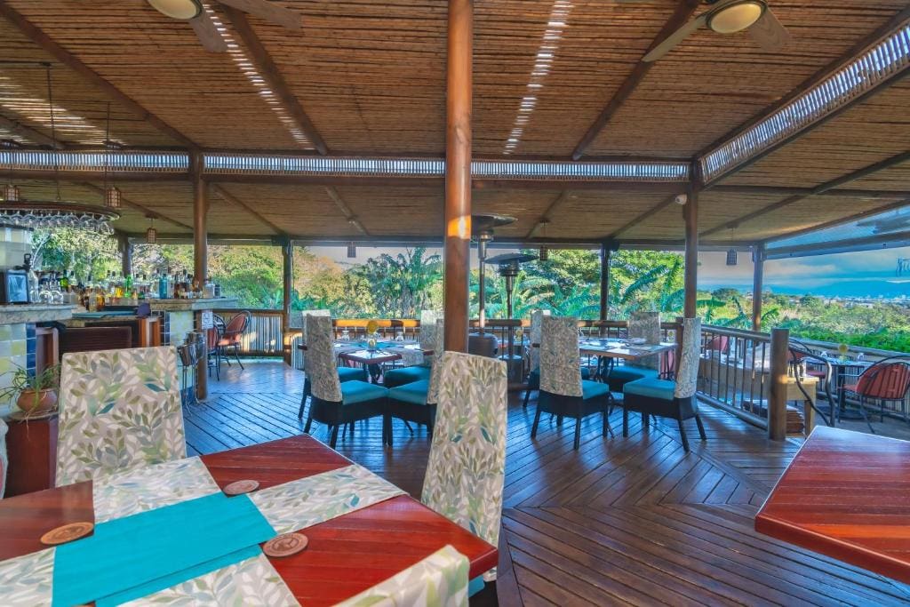 Open-air restaurant with wooden floors and ceiling, colorful cushioned chairs, and tables with leaf-patterned covers. The space overlooks lush greenery and distant ocean views, creating a relaxed, tropical atmosphere.
