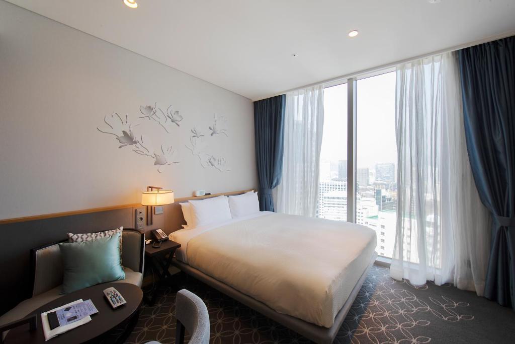 A modern hotel room with a large bed, white bedding, a small round table with a chair, wall art, and floor-to-ceiling windows with blue and white curtains, overlooking a cityscape.