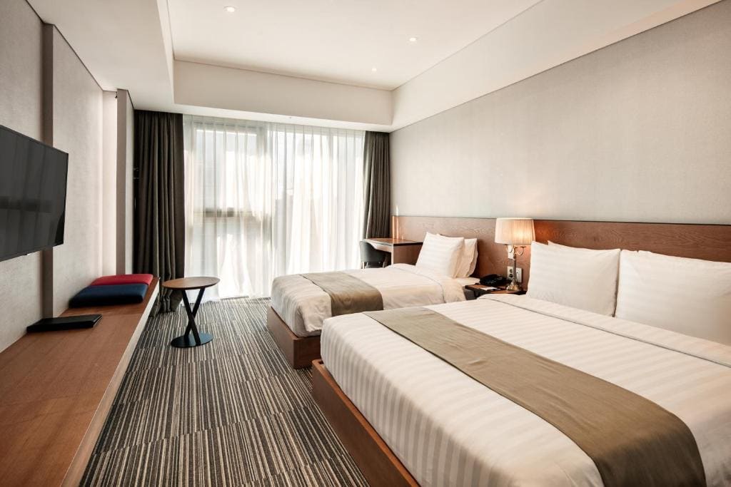 Modern hotel room with two double beds, white bedding, a bedside lamp, a wall-mounted TV, a desk by the window, floor-to-ceiling curtains, and neutral-toned decor. Natural light fills the space.