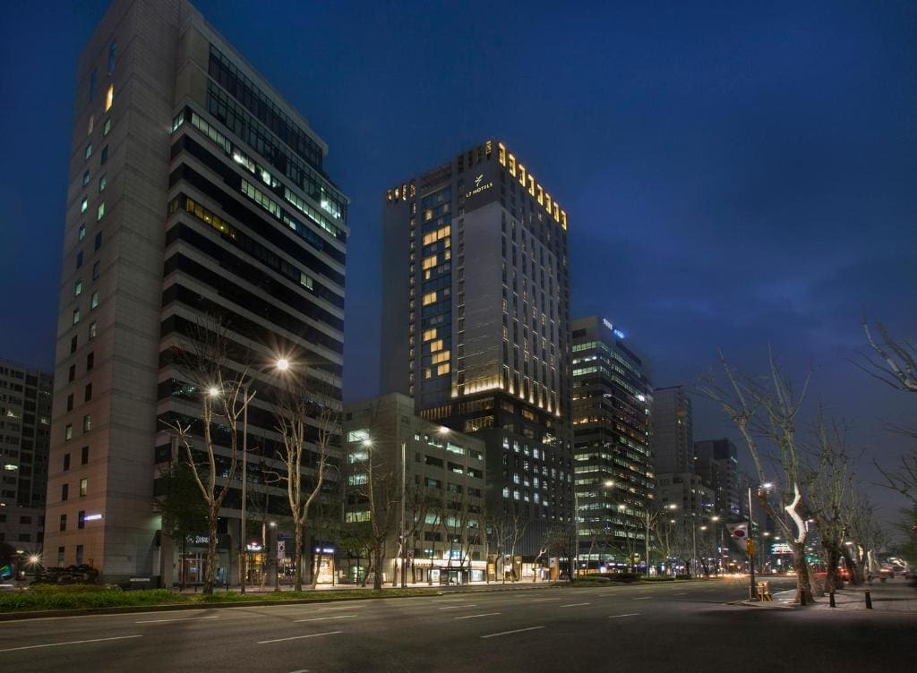 A wide city street lined with tall modern office buildings at night, illuminated by streetlights and building lights, with a few sparse leafless trees along the sidewalk.