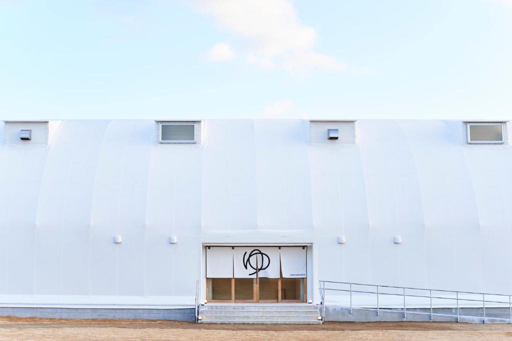 A minimalist white building with a curved facade, small rectangular windows, and a centered entrance. The door features a circular black symbol, and a ramp leads up to the entrance. The sky is clear and blue.