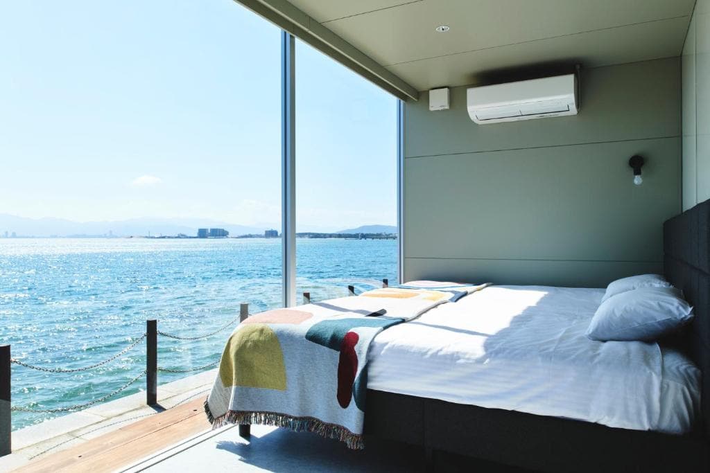 A modern bedroom with large floor-to-ceiling windows overlooks a bright blue sea. Sunlight fills the room, illuminating a neatly made bed with a colorful blanket and pillows. An air conditioner is mounted on the wall.