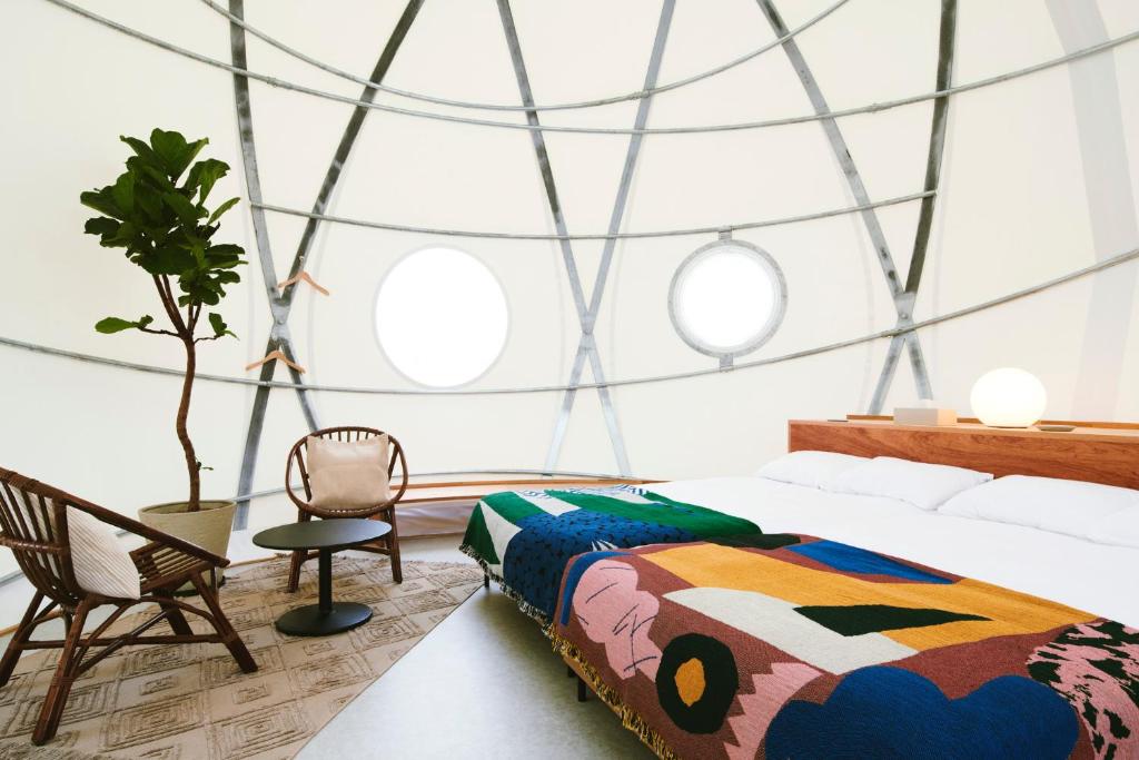 A modern dome-shaped room with circular windows, a colorful patterned blanket on a bed, wooden chairs, a small round table, a potted plant, and soft, diffused lighting.