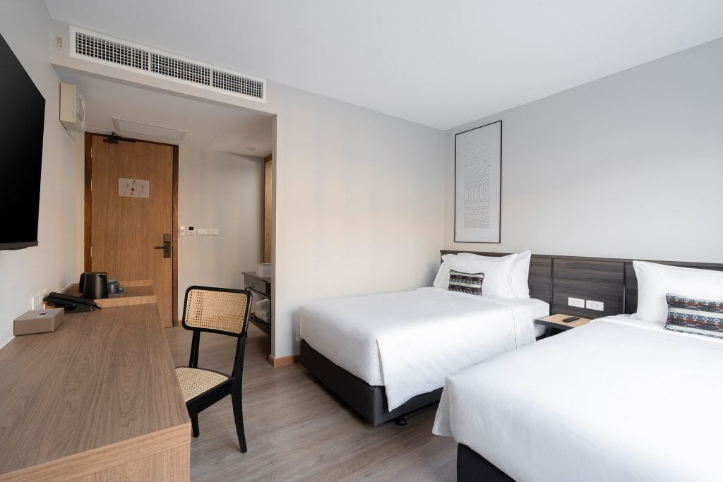 A modern hotel room with two single beds, a wooden desk with a chair, a wall-mounted TV, and a door. The décor is minimal, with white walls and light wood flooring. A framed picture hangs above the beds.
