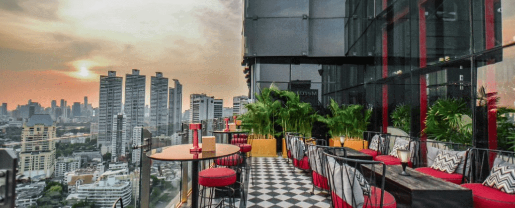A rooftop terrace with red chairs, patterned cushions, and tables overlooks a city skyline at sunset. Potted plants line the edge, and tall buildings are visible in the background under a colorful sky.