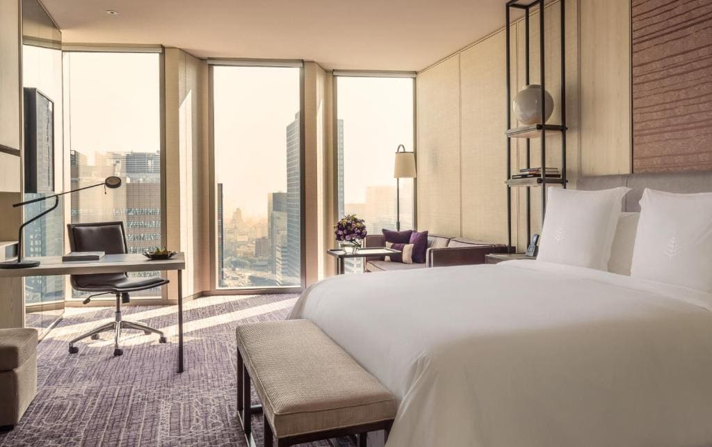 A modern hotel room with a large bed, a work desk and chair by tall windows overlooking a cityscape, a small bench at the bed’s end, and a cozy sitting area with a sofa and decorative lamp.