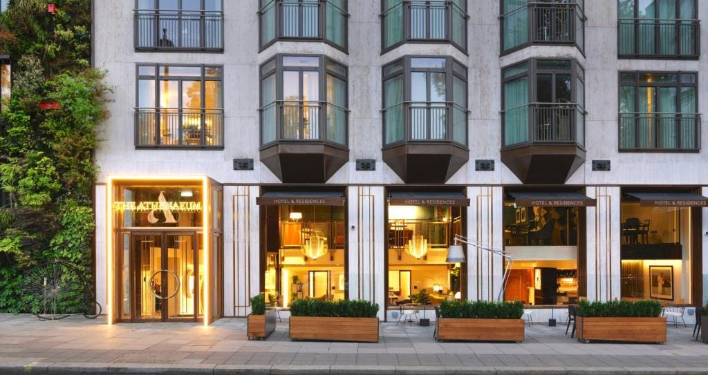 Exterior view of a modern hotel and residences building with large windows, a well-lit entrance, and potted plants along the sidewalk; greenery is visible on the wall to the left.