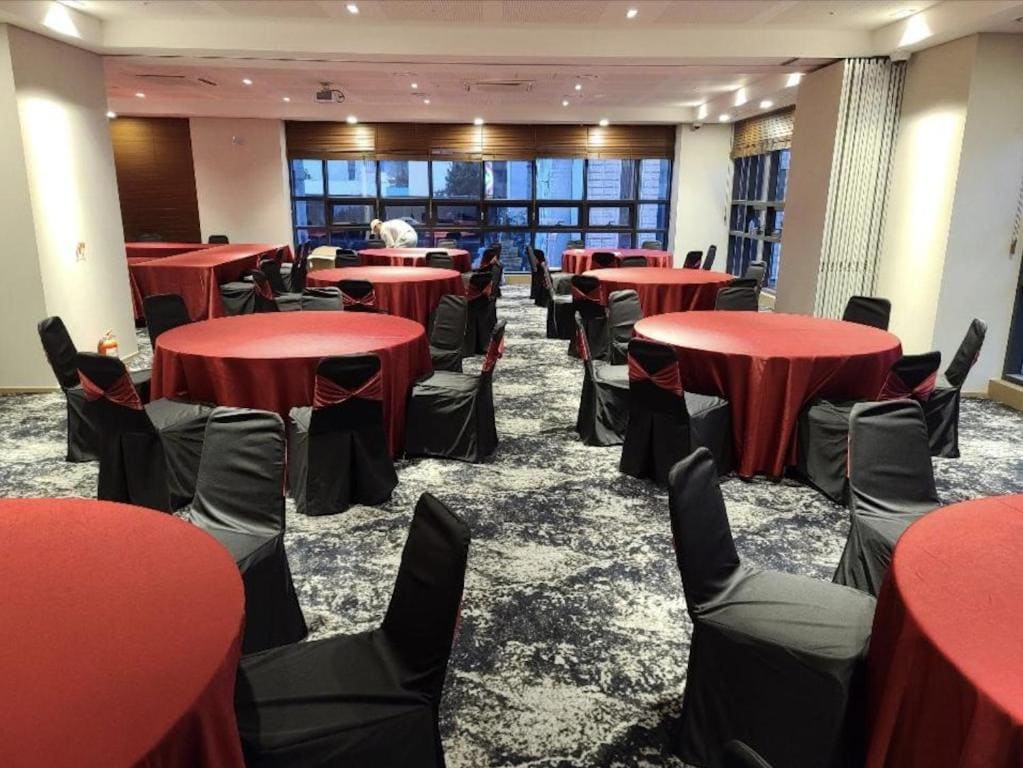 A banquet hall with round tables covered in red tablecloths and black chair covers. The room has large windows, patterned carpet, and a person arranging one of the tables in the background.