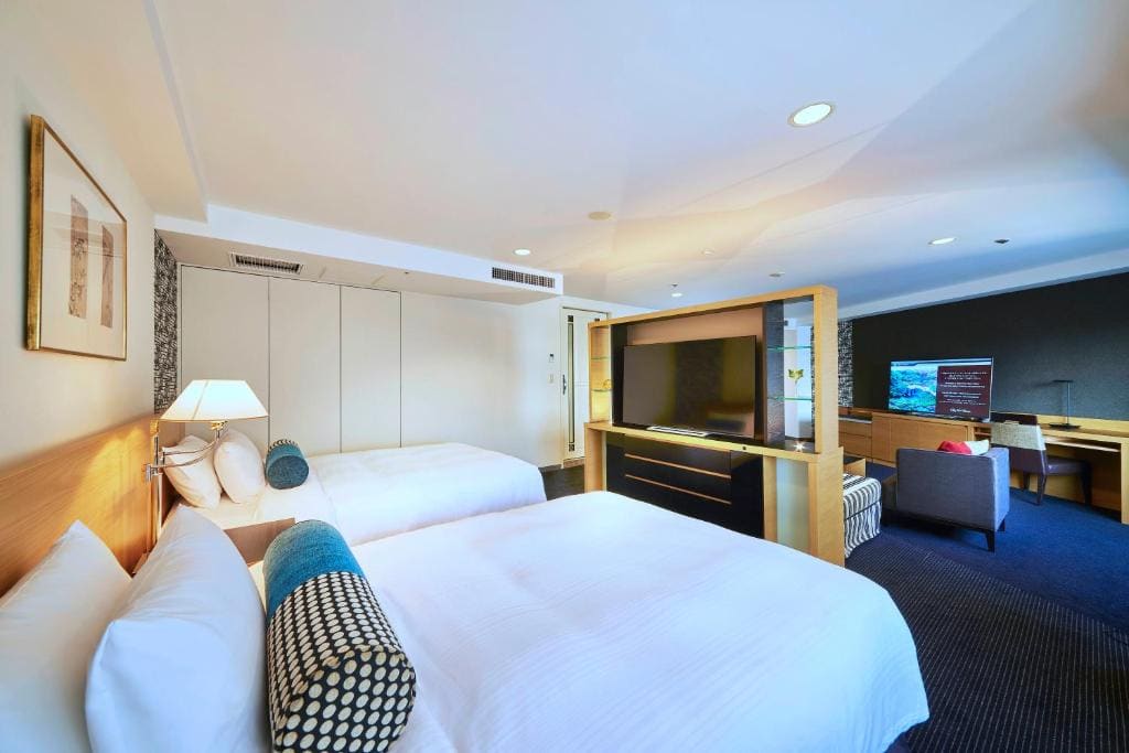 A spacious hotel room with two large beds, white bedding, a bedside lamp, a wall-mounted TV on a divider, a desk with a chair, and a lounge chair. The room is well-lit with modern decor.