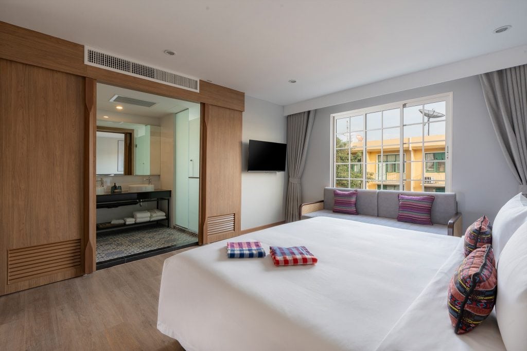 A modern hotel room with a large bed, colorful pillows, a wall-mounted TV, a sofa by a window with curtains, and an open bathroom area with a mirror and sink. Towels are neatly placed on the bed.