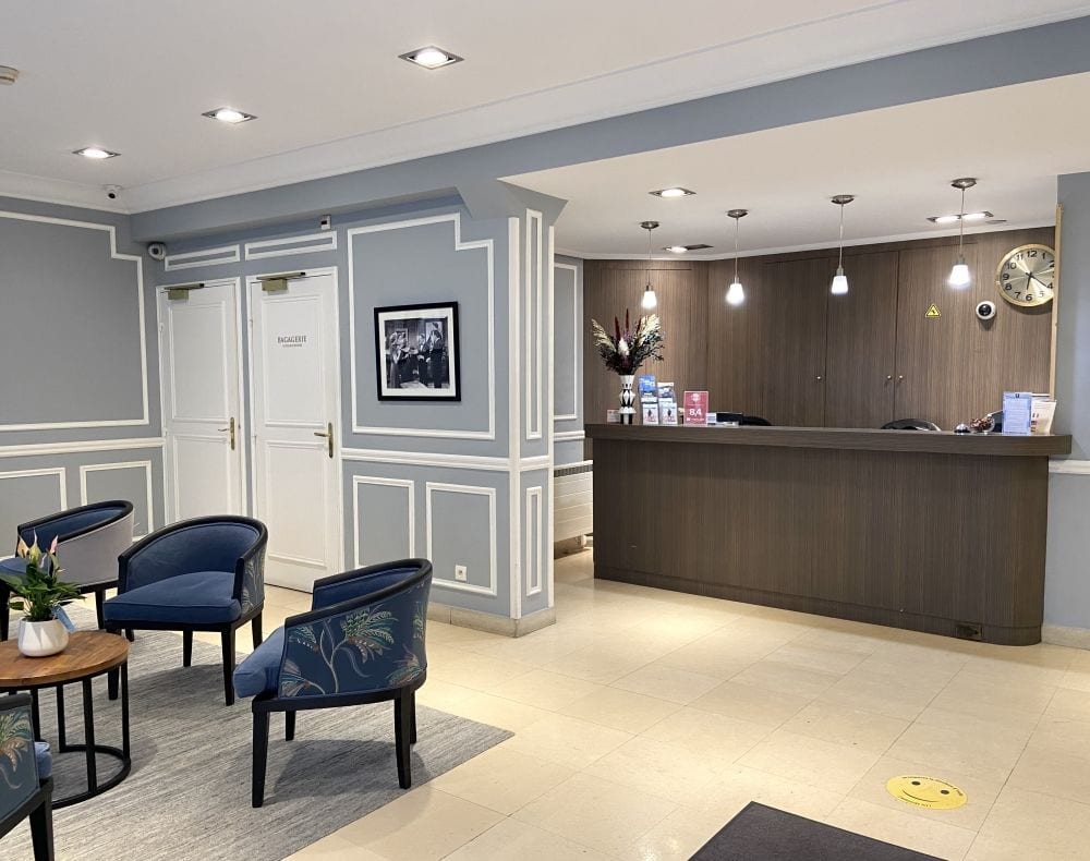 Modern hotel lobby with blue and gray decor, a wooden reception desk with hanging lights, blue upholstered chairs, a small round table with plants, and framed art on the wall. Double doors are visible in the background.