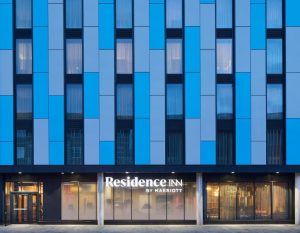 The exterior of a modern hotel building with a blue and gray paneled facade, vertical windows, and a lit sign reading Residence Inn by Marriott above the entrance.
