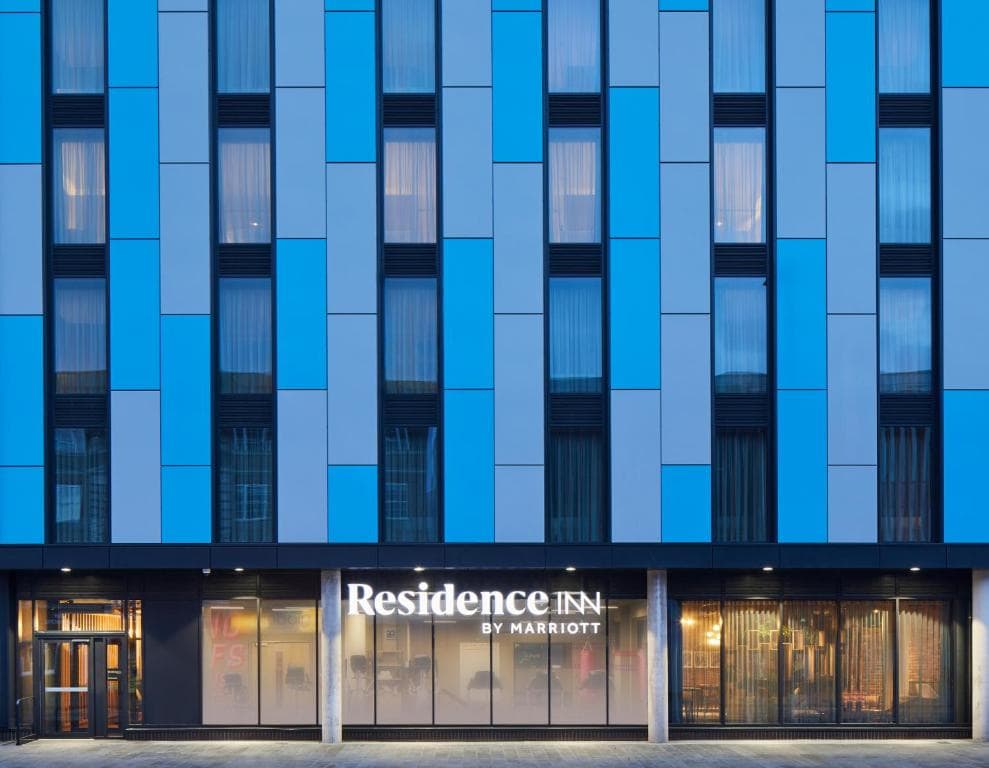 The exterior of a modern hotel building with a blue and gray paneled facade, vertical windows, and a lit sign reading Residence Inn by Marriott above the entrance.