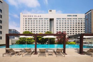 Outdoor swimming pool with lounge chairs and umbrellas in front of a modern hotel building labeled Maison Glad, surrounded by greenery and clear blue sky.