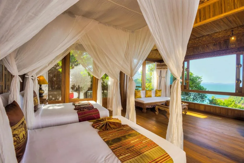 A tropical bedroom with two canopy beds draped in white fabric, wooden floors, and large windows offering views of lush greenery and the ocean. Sunlight fills the cozy, elegant space decorated with warm, earthy tones.