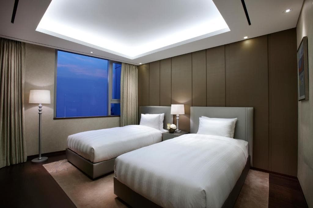 Modern hotel room with two single beds, white bedding, a bedside table with a flower vase, a floor lamp, curtains, and a window showing a blue evening sky. The room has soft lighting and a minimalist design.