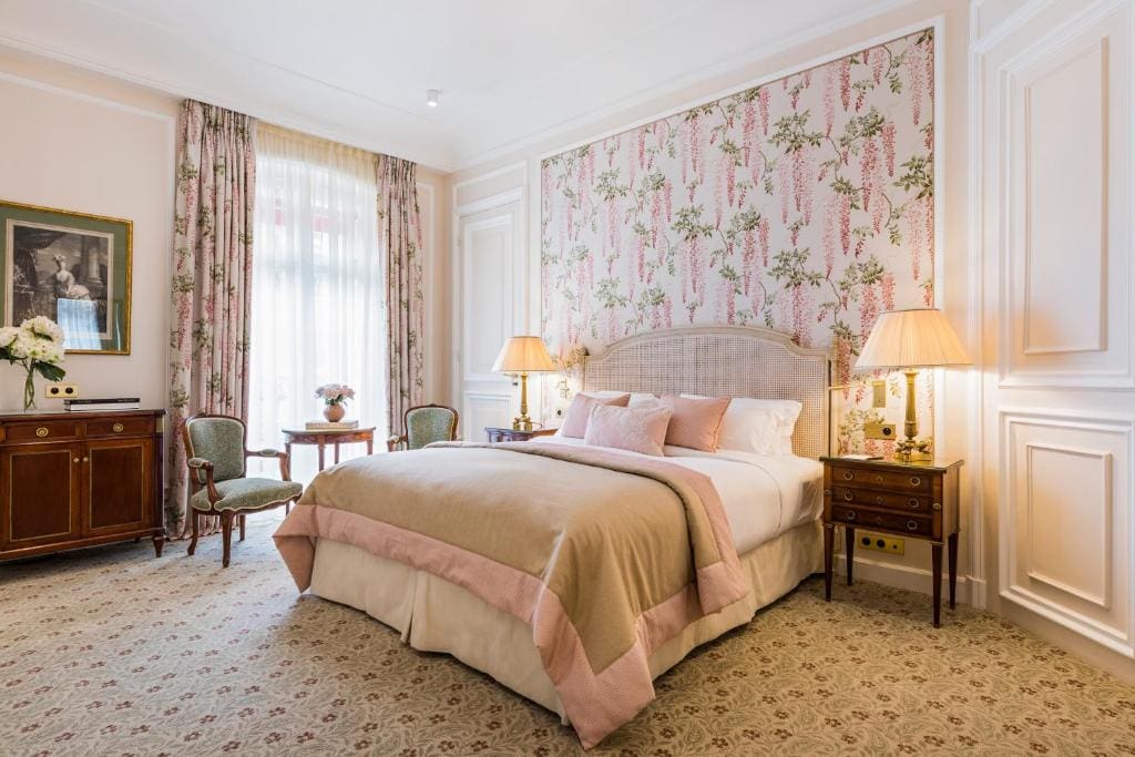 Elegant hotel bedroom with a large bed, pink and beige bedding, floral wallpaper, vintage nightstands with lamps, two green chairs, a small round table by the window, and classic decor. Natural light fills the room.