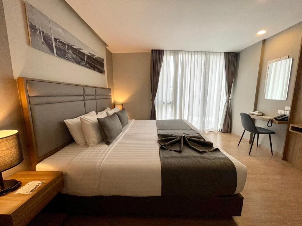 A modern hotel room with a large neatly made bed, two bedside tables with lamps, a desk and chair, a mirror, and floor-to-ceiling windows covered by sheer curtains and dark drapes.