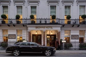 A black luxury car is parked in front of the entrance to Browns Hotel, a classic white building with large windows, potted plants, and a doorman standing by the door.