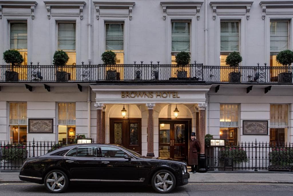 A black luxury car is parked in front of the entrance to Browns Hotel, a classic white building with large windows, potted plants, and a doorman standing by the door.